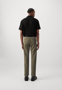 Chemise noire à manches courtes avec un tissage ouvert, associée à un pantalon vert olive et des chaussures noires, vue de dos sur un fond uni.