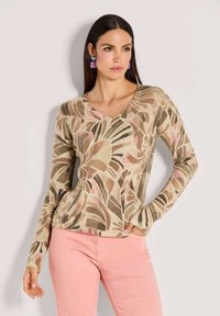 Frau trägt ein langärmliges oberteil in Beige, Pink und Braun mit abstraktem Muster, dazu rosa Hosen und lila Ohrringe, steht mit einer Hand in der Tasche.