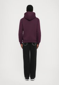 Άνδρας γυρισμένος προς τα πίσω, φορώντας μια βυσσινί hoodie, μαύρα παντελόνια σε φαρδιά γραμμή με ραμμένα πίσω τσέπες και μαύρα με λευκά sneakers, σε λευκό φόντο.