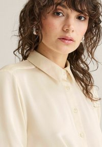 Ung kvinde med bølget brunt hår iført en cremefarvet skjorte med knapper og klassisk krave, vendt let mod venstre.