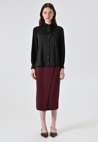 Schwarze Bluse mit langen Ärmeln und einer Schleifen-Detail, kombiniert mit einem bordeauxfarbenen Bleistiftrock. Das Model trägt bordeauxfarbene High Heels. Glatte Stofftextur.