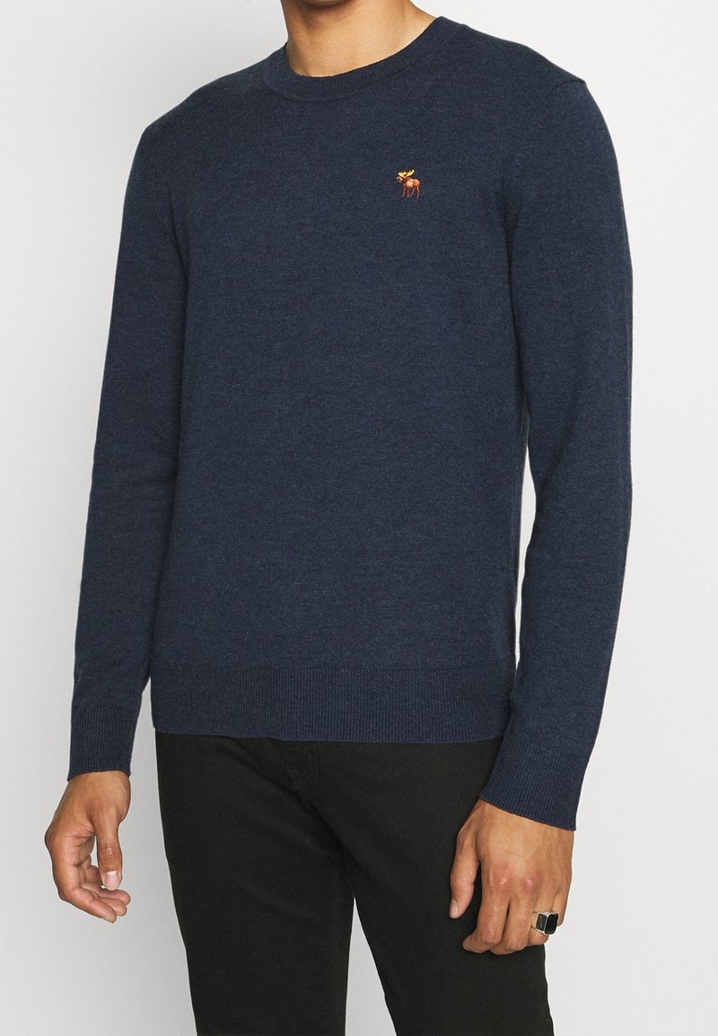 Homme portant un pull bleu foncé à manches longues avec un petit logo élan orange sur la poitrine, assorti à un pantalon noir.