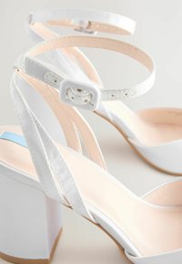 Sandali bianchi con tacco alto e spesso, dal design con cinturini incrociati. Texture liscia con una soletta di un rosa chiaro in contrasto.