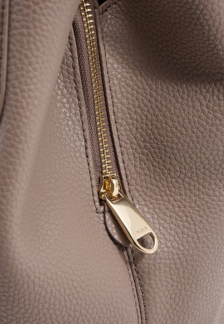 Widok z góry na detal zamka błyskawicznego w beżowej torebce ze skórzanym wykończeniem. Obejmuje złocony uchwyt zamka z subtelnym logotypem, podkreślającym kunszt wykonania.