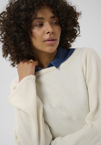 Pull en tricot de couleur crème avec des manches larges, porté sur une chemise en denim foncé. Texture douce, design minimaliste et coupe décontractée.