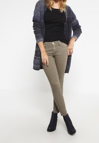 Dunne olijfgroene jeans gecombineerd met een zwarte top en een lange, getextureerde marineblauwe cardigan. Draag met zwarte enkellaarsjes.