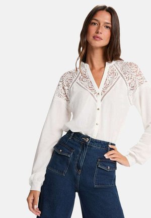 Mujer con blusa blanca de manga larga con detalles de encaje en los hombros y jeans oscuros de talle alto con grandes bolsillos delanteros, mano en la cadera.