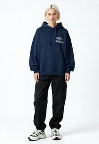 Felpa con cappuccio blu navy con tasca frontale, con il testo bianco "HÔTEL DE L’AMOUR." Abbinata a pantaloni cargo neri e sneakers bianche.