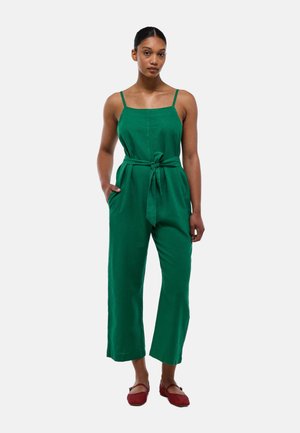 Jumpsuit en lin vert avec des bretelles réglables, une ceinture à nouer à la taille, des jambes larges et des poches latérales. Porté avec des chaussures plates rouges.