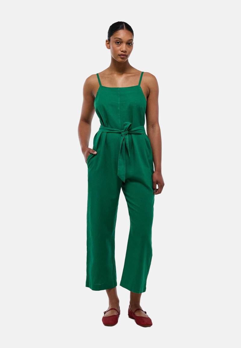 Jumpsuit en lin vert avec des bretelles réglables, une ceinture à nouer à la taille, des jambes larges et des poches latérales. Porté avec des chaussures plates rouges.