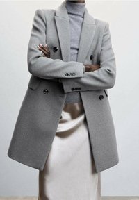 Grå uldblandet dobbeltradet frakke med sorte knapper, strukturerede klapper og sidelommer, lagdelt over en lys grå turtleneck og en satin-nederdel.