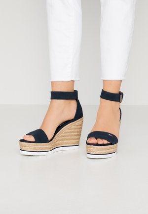 Marineblå kilehæle med espadrille-lignende jute-detaljer, åben tå og ankelrem. Hvid sål med blød, ruskindslignende tekstur.