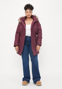 Parka burgundy com capuz de pêlo sintético, fecho frontal com zipper e fechos de pressão. Combinada com jeans azuis de cintura alta e sapatos slip-on bege.
