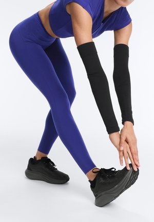 Personne portant des leggings bleus et des chauffe-bras noirs s'étirant en touchant une chaussure noire avec les mains.