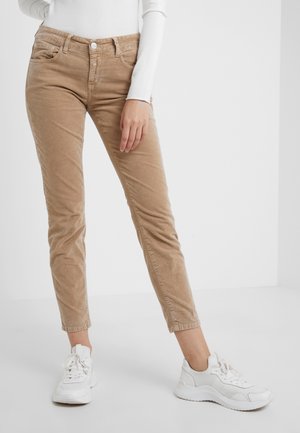 Femme portant un pantalon slim en velours côtelé beige, un haut blanc à manches longues et des baskets blanches, debout sur un sol blanc contre un mur blanc.