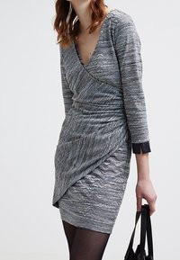 Šedé textura šaty s volným střihem a V výstřihem, dlouhými rukávy a nařaseným předním dílem. Akcentováno černými manžetami, doplněno černou kabelkou.