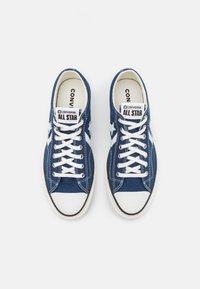Blue canvas sneakers with white rubber toe cap and sole. Features white laces and star logo. Labels include "Converse" and "All Star."