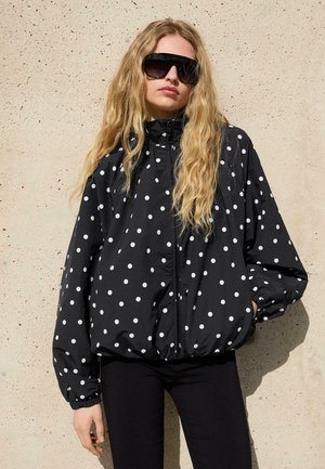 Ženska z dolgimi blond lasmi, ki nosi velike črne sončna očala in črno jakno z belimi pikami, stoji ob bež steni.