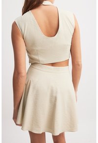 Vaalean beige kahdesta osasta koostuva asu, joka sisältää lyhyen yläosan selkäaukiolla ja levenevät hameen, valmistettu teksturoidusta kankaasta.