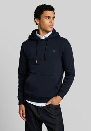 Junger Mann, der einen dunkelblauen Hoodie über einem weißen Hemd mit Knöpfen trägt, steht mit einer Hand in der Tasche und schaut seitlich vor einem schlichten Hintergrund.