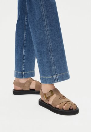 Sandalias de ante marrón con una suela negra gruesa, con una correa en el tobillo y una hebilla dorada. Usadas con vaqueros anchos de pierna en azul claro.