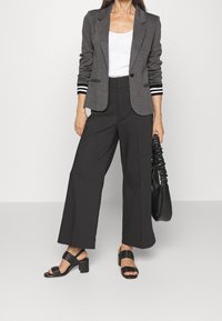 Blazer gris avec poignets rayés, débardeur blanc, pantalon noir à jambes larges et sandales noires à talons. Le modèle tient un sac à main noir.
