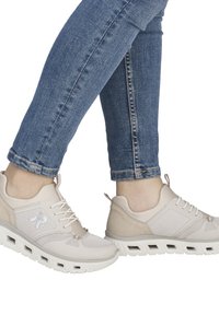 Beżowe buty sportowe z siateczką i materiałami syntetycznymi. Posiadają sznurowadła, oznaczenie marki z boku oraz amortyzowaną podeszwę. Noszone z niebieskimi dżinsami.