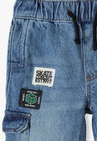 Denim cargo pants in light blue with an elastic waistband, black drawstring, and two patches featuring text and logo details.