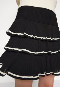 Černá skládací sukně se třemi patry, zdobená bílým volánkem. Materiál má texturovaný povrch s horizontálními proužky pro tvar.