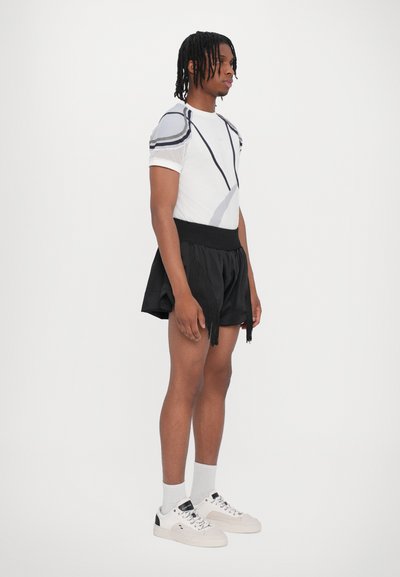 Weißes Netz-T-Shirt mit grauen grafischen Linien und schwarzen Baumwollshorts mit Fransendetail. Getragen mit weißen Sneakers und grauen Socken.