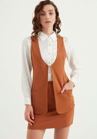 Brun väst med djup V-ringning, två framfickor; bärs över en vit skjorta med krage. Kjol i matchande brunt tyg.