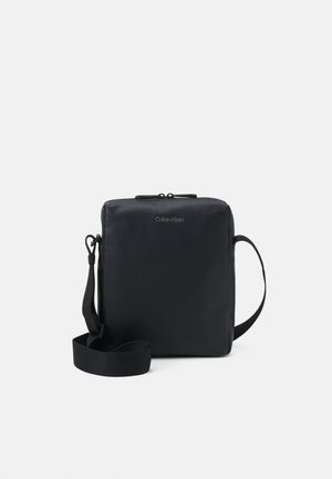 Sac bandoulière rectangulaire noir Calvin Klein avec sangle ajustable et fermeture éclair sur fond blanc.