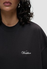 Mustat ylisuuret hupparit, joissa on ribbikuvioitu pyöreä pääntie ja valkoinen brodeerattu "Wrstbhvr" -logo rinnassa. Pehmeä kangas, rento istuvuus.
