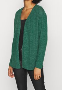 Zielony, otwarty kardigan z miękkiego, fakturowanego materiału. Posiada długie rękawy i luźny krój, nałożony na czarną bluzkę i spodnie.