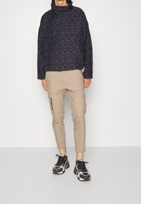 Vadderad marinblå pull-over med hög krage och geometriskt mönster, kombinerad med beiga cargobyxor med sidofickor och svarta sneakers.