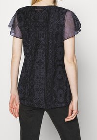 Mujer vestida con una blusa negra de patrón que tiene mangas cortas transparentes y onduladas, de pie con las manos relajadas a los lados, mirando hacia lejos de la cámara.