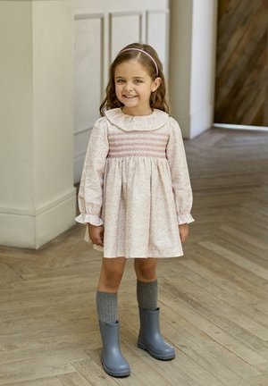 Vestido rosa claro con mangas largas y un cuello con volantes; cuerpo fruncido con patrón floral. Medias gris de rodilla y botas de goma grises.