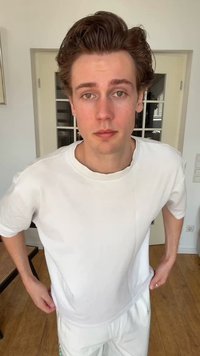 Νεαρός άνδρας με ανοιχτόχρωμο δέρμα και καστανά μαλλιά, που φοράει λευκό μπλουζάκι και λευκό παντελόνι, στέκεται μέσα σε κλειστό χώρο με τα χέρια στη μέση, κοιτάζοντας μπροστά.