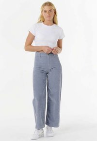 Hvid, ribbet T-shirt kombineret med højtaljede, blå bukser med lodrette striber og brede ben samt hvide sneakers, hvilket skaber et afslappet outfit.