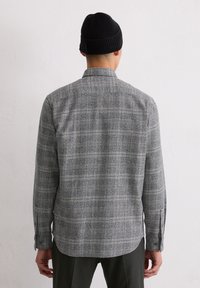 Grijze ruitjeshemd met lange mouwen, een kraag en zijslitsen, gemaakt van textuurstof. Draagt een zwarte beanie.