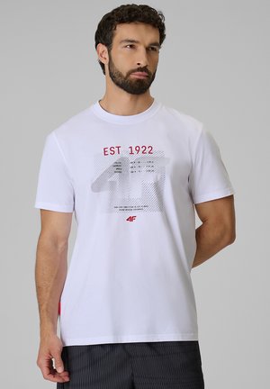 Mężczyzna z brodą w białej koszulce z czerwonym napisem "EST 1922" i szarą grafiką, stojący na jednolitym jasnoszarym tle.