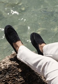 Svarta mockaloffers med en slät yta och sömmar som detaljer, placerade på en sten nära klart vatten, med vita byxor nedanför.