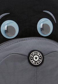 Kipling rugzak met een zwarte stoffen body, een grijze ritsvak, grote blauwe plastic ogen en een cirkelvormig logo op een textuur patch.