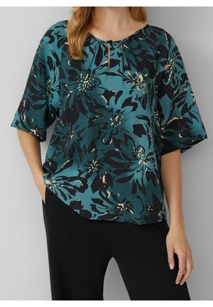 Mujer con blusa azul verdoso con estampado floral negro y beige y pantalones negros, de pie con una mano en el bolsillo.