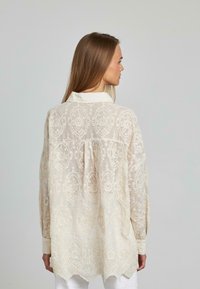 Helle beige Bluse mit floraler Spitze, langen Ärmeln und weißem Kragen. Verfügt über einen gewellten Saum und filigrane Stickdetails.