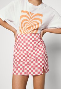 T-shirt branca com um padrão de corações laranja, combinado com uma saia mini xadrez nas cores rosa e branca, com detalhes florais e uma fenda lateral.