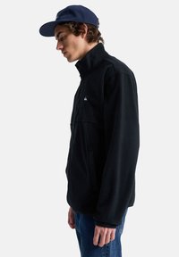 Giacca in pile nera con colletto alto, zip frontale, tasche laterali e piccolo logo. Indossata con un cappellino blu navy e jeans blu.