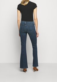 Jeans de campana de mezclilla en un tono oscuro con cinco bolsillos y presillas para cinturón, combinados con una camiseta negra ajustada; textura suave. Zapatos de tacón marrones.