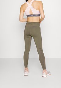 Sottogiacca sportiva rosa con design a croce e banda nera con logo abbinata a leggings verde oliva. Il modello indossa scarpe da ginnastica di colore chiaro.