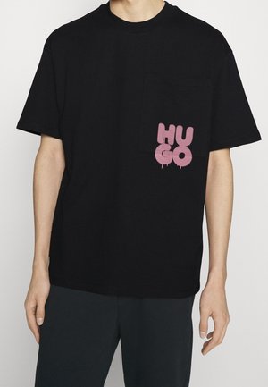 Mężczyzna ubrany w czarną koszulkę z kieszenią na piersi z różowymi graffiti w stylu "HUGO" oraz czarne spodnie.